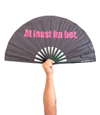 A person holding a dark gray folding hand fan with pink text that reads 'At least im hot.' 
