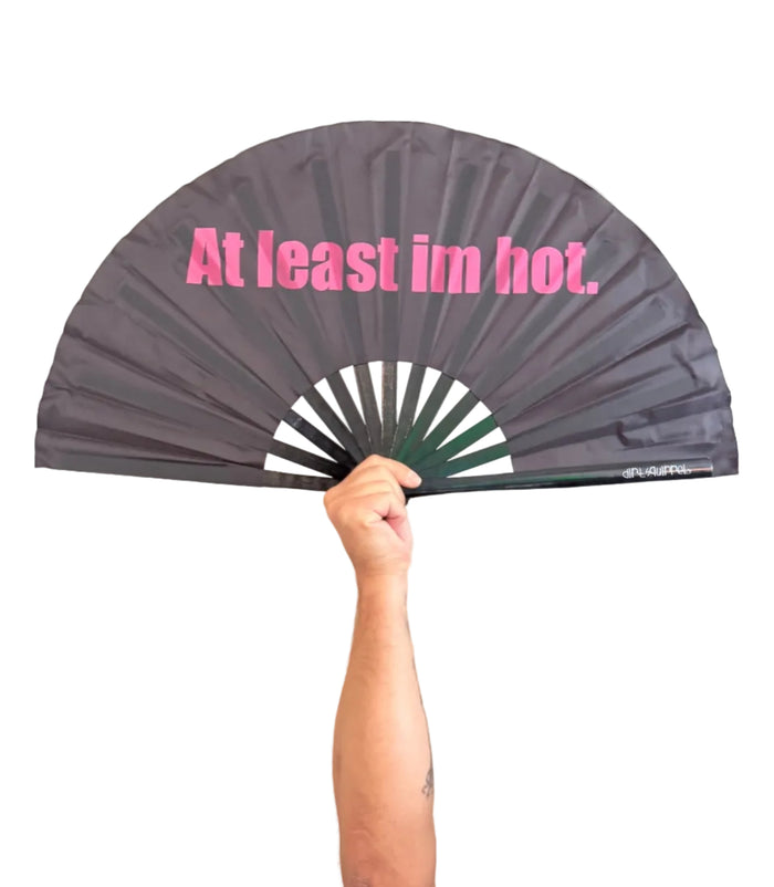 A person holding a dark gray folding hand fan with pink text that reads 'At least im hot.' 