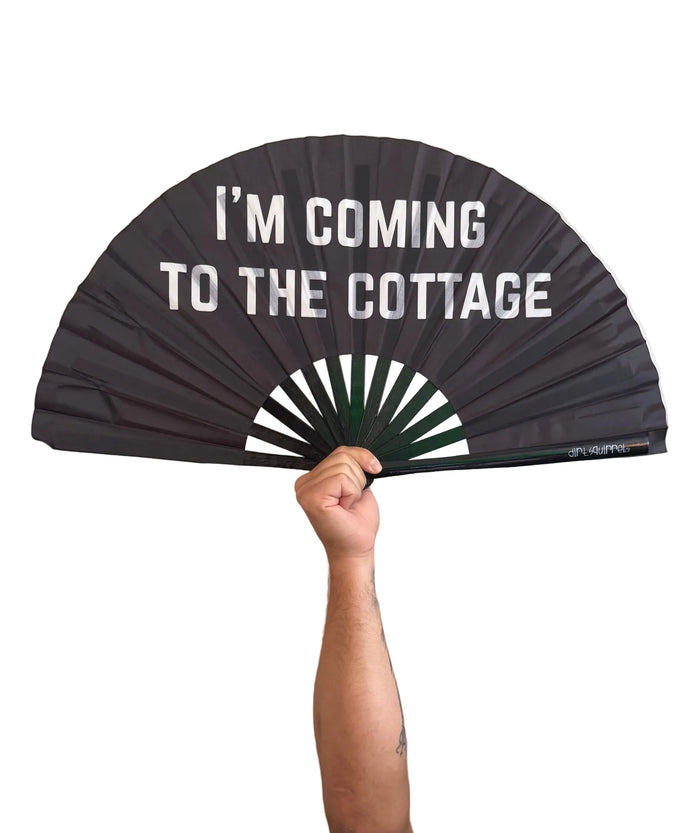 A black folding hand fan with white text reading 'I'M COMING TO THE COTTAGE' and a white-and-green geometric center, held by a person in a black graphic t-shirt in an indoor setting.